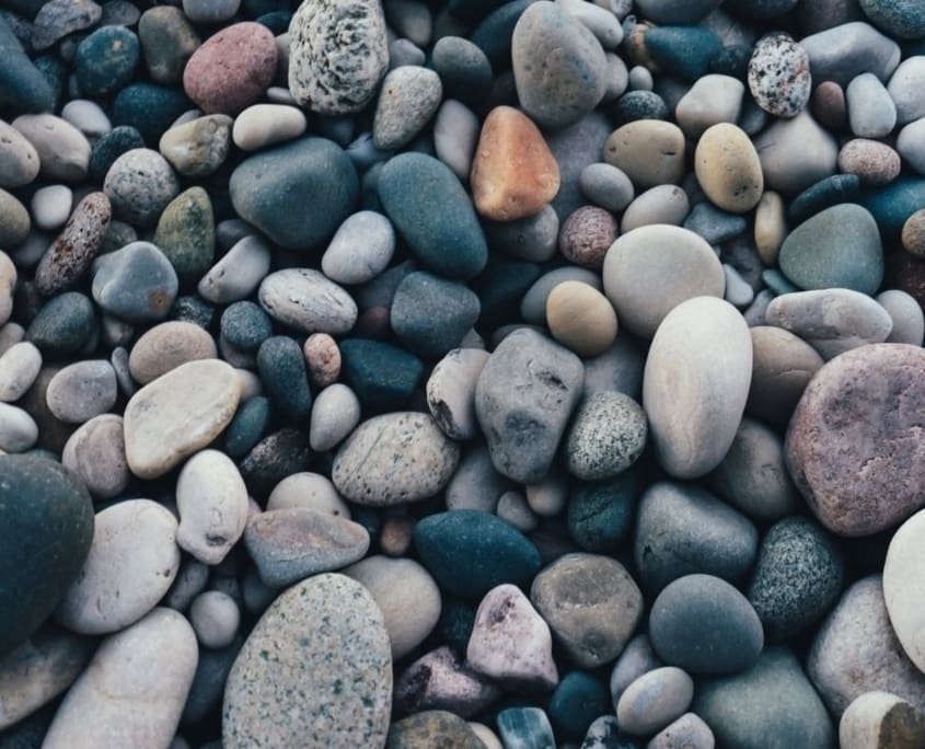 Landscape Design Brisbane Close-up of a collection of smooth, multi-colored pebbles and stones of varying sizes. The stones range from dark gray and blue to lighter shades of tan, ivory, and a few in muted orange and green. They are tightly packed together, covering the entire frame. Live Outdoors