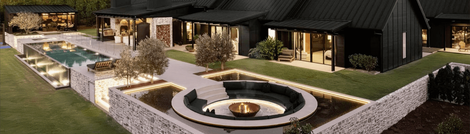 Landscape Design Brisbane A modern, luxurious house with black exterior, featuring a large outdoor area with a swimming pool, sunken circular fire pit with seating, and landscaped gardens. The house has large glass windows and stone walls, set against a backdrop of trees. Live Outdoors