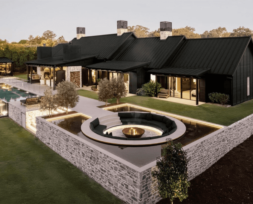 Landscape Design Brisbane A modern, luxurious house with black exterior, featuring a large outdoor area with a swimming pool, sunken circular fire pit with seating, and landscaped gardens. The house has large glass windows and stone walls, set against a backdrop of trees. Live Outdoors