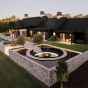 Landscape Design Brisbane Modern luxury home with black exterior, surrounded by greenery. Features a long pool, wooden deck, and sunken fire pit area. Elegant outdoor lighting highlights the architectural design. Split-level layout and large windows enhance the contemporary aesthetic. Live Outdoors