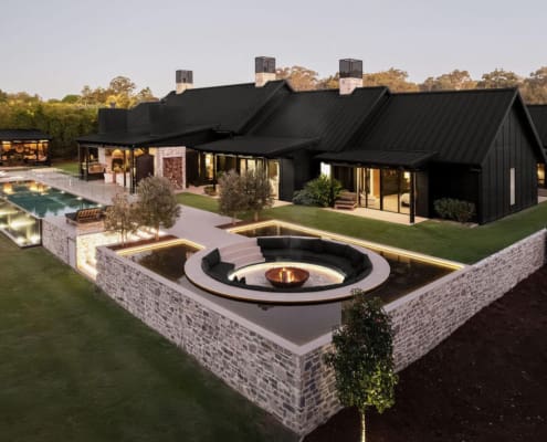 Landscape Design Brisbane Modern luxury home with black exterior, surrounded by greenery. Features a long pool, wooden deck, and sunken fire pit area. Elegant outdoor lighting highlights the architectural design. Split-level layout and large windows enhance the contemporary aesthetic. Live Outdoors