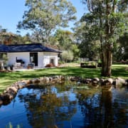 Landscape Design Brisbane A serene backyard features a white house with black trim, surrounded by lush green lawn and tall trees. A small pond bordered by rocks reflects the clear blue sky and trees, adding tranquility to the landscape. Outdoor seating is visible near the house. Live Outdoors