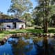 Landscape Design Brisbane A serene backyard features a white house with black trim, surrounded by lush green lawn and tall trees. A small pond bordered by rocks reflects the clear blue sky and trees, adding tranquility to the landscape. Outdoor seating is visible near the house. Live Outdoors