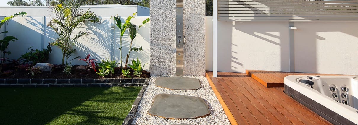 Landscape Design Brisbane Outdoor patio area featuring a wooden deck with an integrated hot tub, a stone path leading to an outdoor shower with a pebble base, and surrounding green artificial grass and tropical plants against a white fence. Live Outdoors