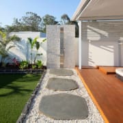 Landscape Design Brisbane Outdoor patio area featuring a wooden deck with an integrated hot tub, a stone path leading to an outdoor shower with a pebble base, and surrounding green artificial grass and tropical plants against a white fence. Live Outdoors