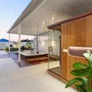 Landscape Design Brisbane Outdoor wellness area featuring a wooden sauna unit and a large jacuzzi, surrounded by white tiled flooring. The space has a modern design with a covered roof, lush green plants, and ambient lighting, creating a relaxing atmosphere. Live Outdoors