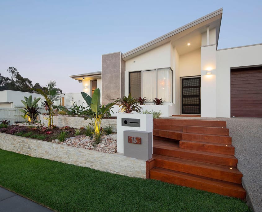 Landscape Design Brisbane Modern single-story house with large windows, a wooden entrance staircase, and a contemporary garden featuring various plants and illuminated landscaping. A gravel driveway leads to a closed garage, and the house number “15” is displayed on a stone marker. Live Outdoors