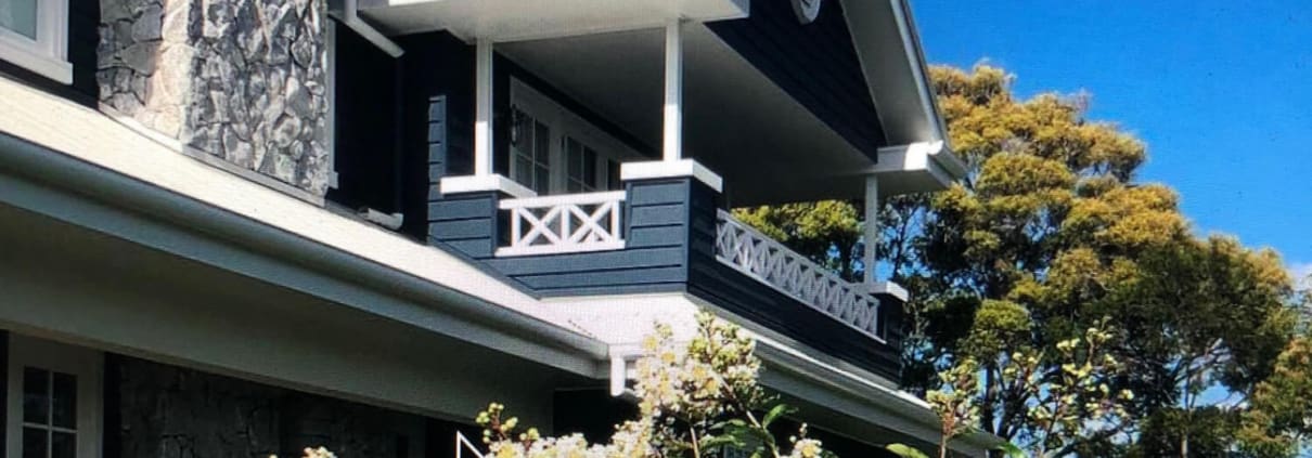 Landscape Design Brisbane A two-story house with dark blue walls, white trim, and a stone facade. There is a small balcony on the upper floor. The front of the house is surrounded by blooming white flowers and trees under a bright blue sky with a few white clouds. Live Outdoors