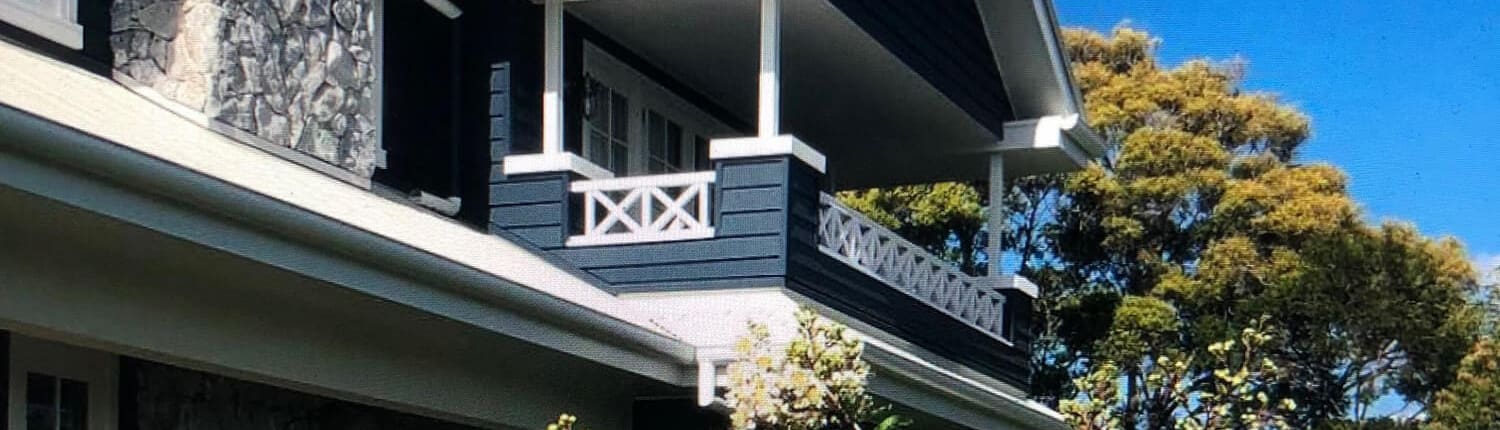 Landscape Design Brisbane A two-story house with dark blue walls, white trim, and a stone facade. There is a small balcony on the upper floor. The front of the house is surrounded by blooming white flowers and trees under a bright blue sky with a few white clouds. Live Outdoors