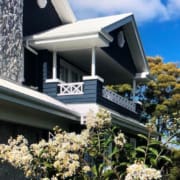 Landscape Design Brisbane A two-story house with dark blue walls, white trim, and a stone facade. There is a small balcony on the upper floor. The front of the house is surrounded by blooming white flowers and trees under a bright blue sky with a few white clouds. Live Outdoors