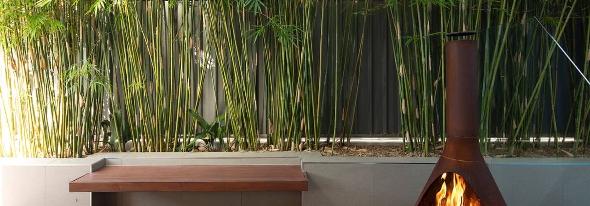 Landscape Design Brisbane A cozy outdoor space with a rust-colored freestanding fireplace, lit and burning brightly. The area includes a wooden bench and a deck, surrounded by a lush bamboo garden and a tall metal fence. The scene is serene and inviting. Live Outdoors