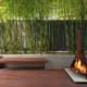 Landscape Design Brisbane A cozy outdoor space with a rust-colored freestanding fireplace, lit and burning brightly. The area includes a wooden bench and a deck, surrounded by a lush bamboo garden and a tall metal fence. The scene is serene and inviting. Live Outdoors