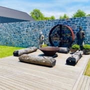 Landscape Design Brisbane A contemporary garden features a wooden deck with rustic log benches, a large fire bowl, and a circular log storage. Two statues are placed near the stone wall at the back, along with trimmed shrubs. There's a modern black-roofed building and trees in the background. Live Outdoors