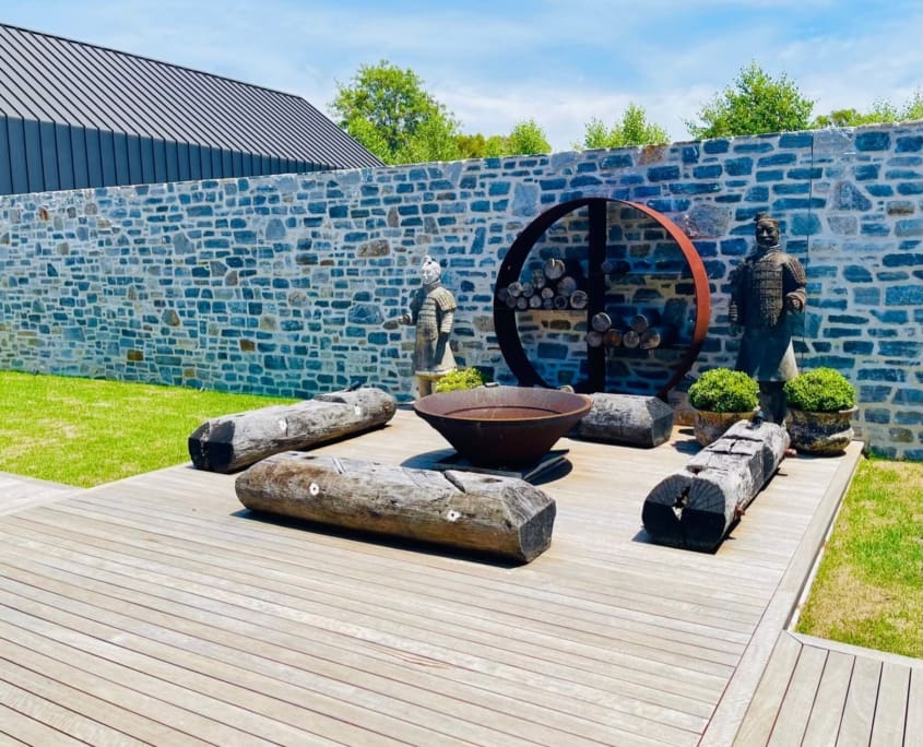 Landscape Design Brisbane A contemporary garden features a wooden deck with rustic log benches, a large fire bowl, and a circular log storage. Two statues are placed near the stone wall at the back, along with trimmed shrubs. There's a modern black-roofed building and trees in the background. Live Outdoors