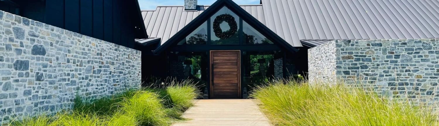 Landscape Design Brisbane A modern building with a stone exterior and metal roof is seen on a sunny day. A wooden walkway leads to the front entrance, which is adorned with a wreath. Tall grass flanks the path on either side, adding a natural touch to the landscape. Live Outdoors