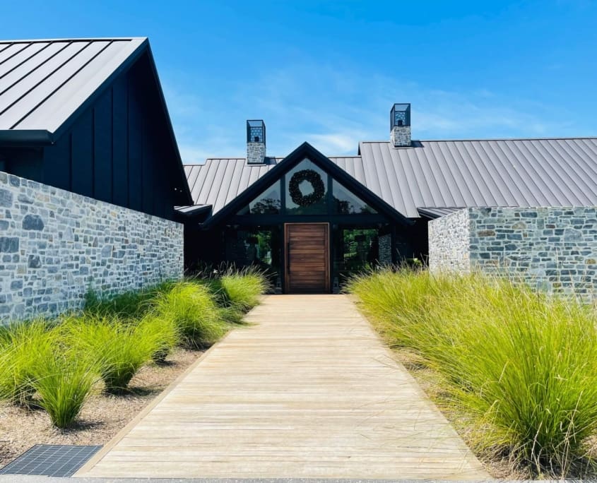 Landscape Design Brisbane A modern building with a stone exterior and metal roof is seen on a sunny day. A wooden walkway leads to the front entrance, which is adorned with a wreath. Tall grass flanks the path on either side, adding a natural touch to the landscape. Live Outdoors