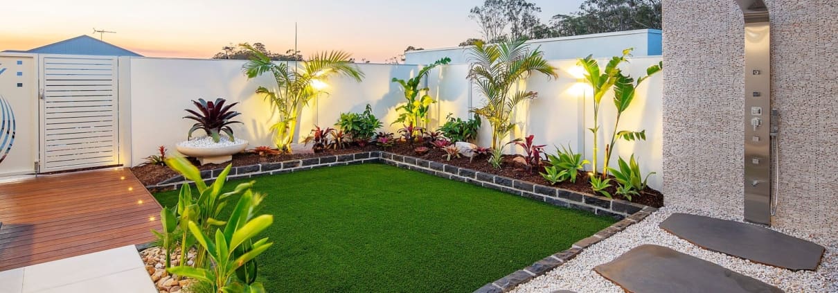 Landscape Design Brisbane A modern backyard featuring a rectangular section of lush green grass surrounded by well-maintained plants and shrubs. There are large stepping stones set in white gravel leading to a contemporary outdoor shower with an adjacent wooden deck area. Live Outdoors