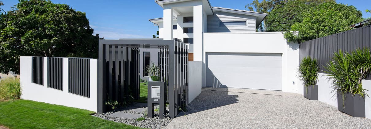 Landscape Design Brisbane A modern, two-story house with a white exterior, flat roof, and large windows. The front yard has a small lawn and gravel driveway leading to a garage. A sleek, black metal gate and fence enclose the property. Potted plants are placed near the entrance. Bright, sunny day. Live Outdoors