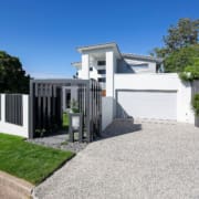 Landscape Design Brisbane A modern, two-story house with a white exterior, flat roof, and large windows. The front yard has a small lawn and gravel driveway leading to a garage. A sleek, black metal gate and fence enclose the property. Potted plants are placed near the entrance. Bright, sunny day. Live Outdoors