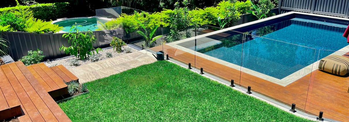 Norman Park Grand and Grander Landscape Design Brisbane A backyard featuring a wooden deck on the left, a green grassy lawn in the center, and a rectangular swimming pool with clear glass fencing on the right. In the background, there are lush plants and trees, with a small elevated round spa to the left of the pool. Live Outdoors