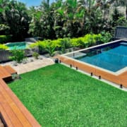 Norman Park Grand and Grander Landscape Design Brisbane A backyard featuring a wooden deck on the left, a green grassy lawn in the center, and a rectangular swimming pool with clear glass fencing on the right. In the background, there are lush plants and trees, with a small elevated round spa to the left of the pool. Live Outdoors