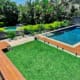 Landscape Design Brisbane A backyard featuring a wooden deck on the left, a green grassy lawn in the center, and a rectangular swimming pool with clear glass fencing on the right. In the background, there are lush plants and trees, with a small elevated round spa to the left of the pool. Live Outdoors