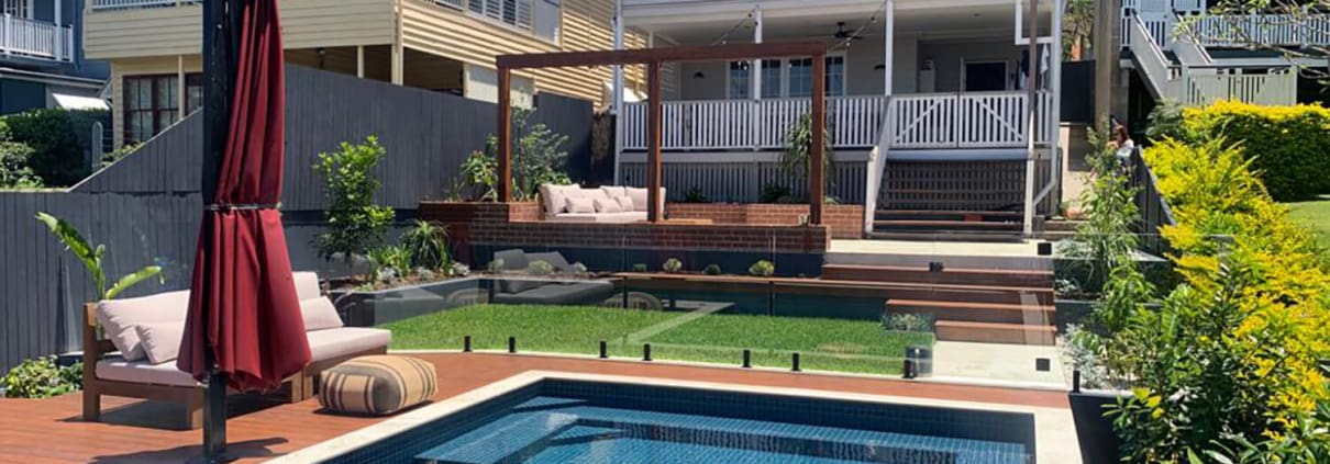 Landscape Design Brisbane A modern backyard features a rectangular pool with blue tiles and an adjacent deck area with a large red umbrella and cushioned seating. The space is landscaped with green grass, plants, and steps leading up to a raised patio area attached to a contemporary two-story house. Live Outdoors