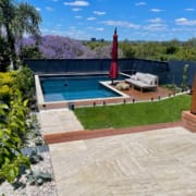 Landscape Design Brisbane A backyard with a rectangular swimming pool surrounded by a wooden deck and glass fence. There's a cushioned lounge chair with a red umbrella alongside the pool, greenery, flowering trees, and a tiled patio in the foreground. The sky is blue with scattered clouds. Live Outdoors