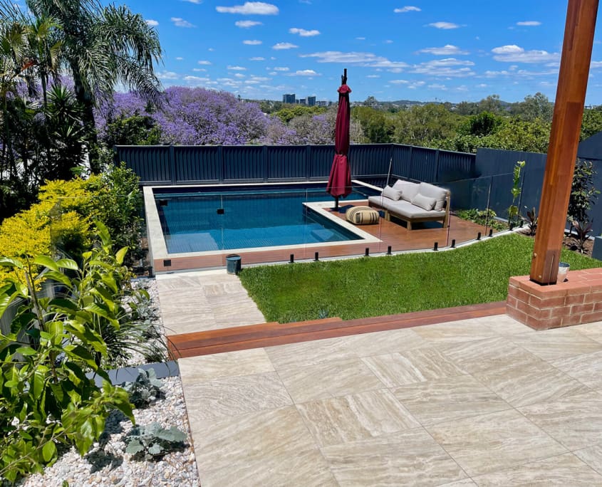 Landscape Design Brisbane A backyard with a rectangular swimming pool surrounded by a wooden deck and glass fence. There's a cushioned lounge chair with a red umbrella alongside the pool, greenery, flowering trees, and a tiled patio in the foreground. The sky is blue with scattered clouds. Live Outdoors