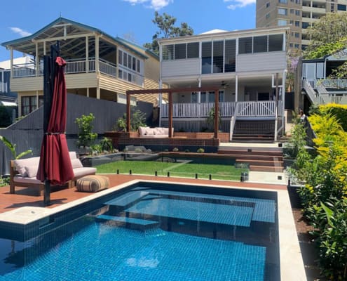 Landscape Design Brisbane A modern backyard features a rectangular pool with blue tiles and an adjacent deck area with a large red umbrella and cushioned seating. The space is landscaped with green grass, plants, and steps leading up to a raised patio area attached to a contemporary two-story house. Live Outdoors