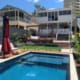 Landscape Design Brisbane A modern backyard features a rectangular pool with blue tiles and an adjacent deck area with a large red umbrella and cushioned seating. The space is landscaped with green grass, plants, and steps leading up to a raised patio area attached to a contemporary two-story house. Live Outdoors