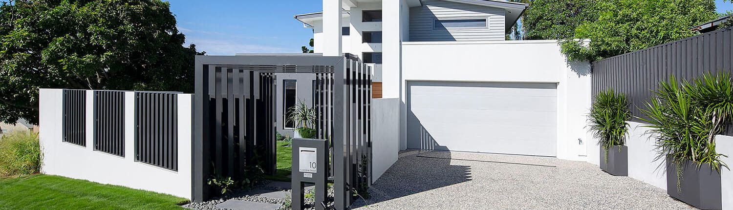 Landscape Design Brisbane Modern white house with a minimalist design, featuring a tall facade and a two-car garage. The entrance includes a gated pathway with black vertical bars, landscaped with green plants and a small lawn. The driveway is made of clean aggregate concrete. Blue sky background. Live Outdoors