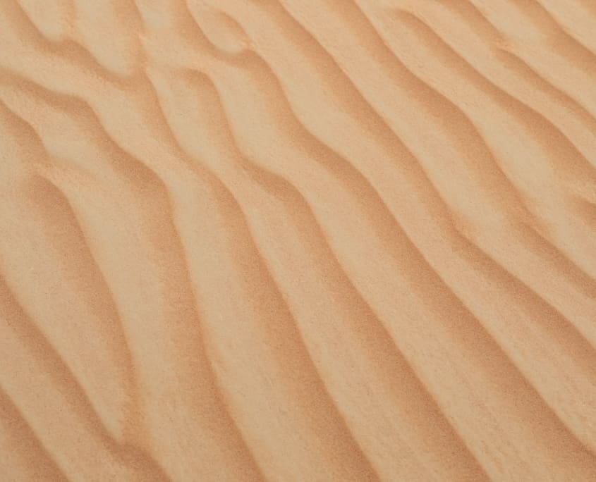 Landscape Design Brisbane A close-up view of a sandy desert surface with a rippling pattern formed by wind. The sand appears golden and smooth, with evenly spaced waves running horizontally across the image. Live Outdoors