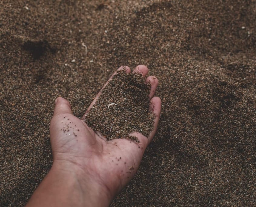 Landscape Design Brisbane A close-up of a hand holding dark, coarse sand, with more sand in the background. The hand is slightly cupped, allowing some grains to spill out. The image highlights the texture and earthy color of the sand. Live Outdoors