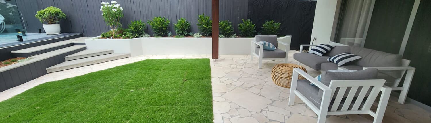 Suburban backyard oasis, Landscape Design Brisbane A backyard with a covered patio featuring white outdoor furniture, including a couch with gray cushions and a striped pillow, and a wicker coffee table. The patio floor is made of light-colored stone, and a well-manicured lawn is adjacent to it. The background shows a wooden fence with plants, a Jacuzzi, and steps leading up to another level of the yard. Live Outdoors