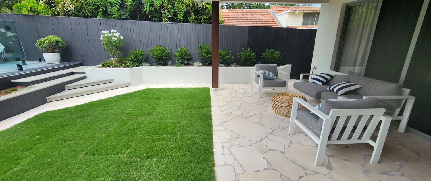 Suburban backyard oasis Suburban backyard oasis, Landscape Design Brisbane A backyard with a covered patio featuring white outdoor furniture, including a couch with gray cushions and a striped pillow, and a wicker coffee table. The patio floor is made of light-colored stone, and a well-manicured lawn is adjacent to it. The background shows a wooden fence with plants, a Jacuzzi, and steps leading up to another level of the yard. Live Outdoors