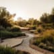 Landscape Design Brisbane A serene garden at sunset features a pebbled pathway winding through lush greenery and various types of plants. Wooden steps lead up to a raised platform. Tall trees frame the peaceful, sun-dappled landscape. Live Outdoors