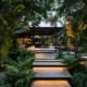 Landscape Design Brisbane A modern outdoor garden with lush green plants, ferns, and large tropical leaves. There are illuminated concrete steps leading to a covered lounging area with chairs and a table. Soft, ambient lighting enhances the serene, nighttime atmosphere. Live Outdoors