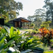 Landscape Design Brisbane A modern, single-story house with large glass windows sits atop a landscaped hill surrounded by lush greenery and tropical plants. Tall trees and vibrant bushes create a serene, natural backdrop under a bright, clear sky. Live Outdoors