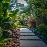 Landscape Design Brisbane A serene garden pathway features square stepping stones surrounded by lush green foliage and various tropical plants. The sun casts dappled light, creating a tranquil and inviting atmosphere along the path. Live Outdoors